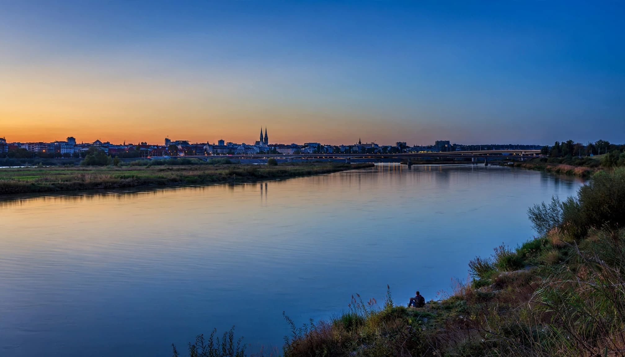A lone photographer watches the Sava River and Zagreb skyline at blue hour.